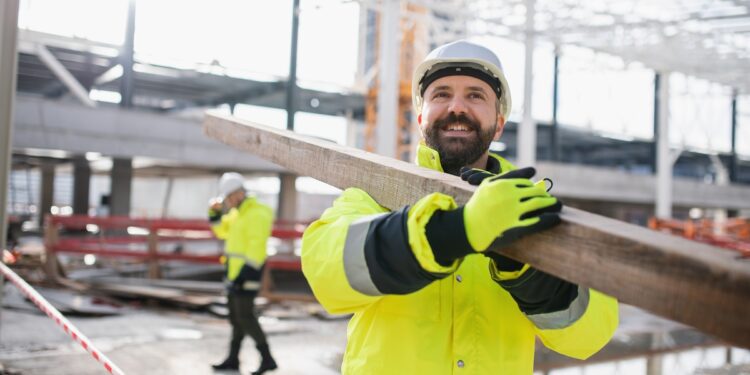 A smiling man wearing a white hard hat, yellow high visibility coat, and yellow high visibility gloves is carrying a long piece of lumber on his shoulder at a construction site.