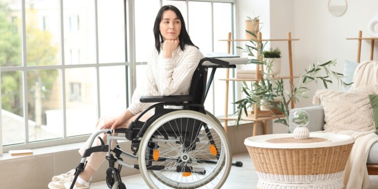 A woman sitting in her wheelchair in front of a large, sunny window. Her head rests on a fist as she looks behind her.