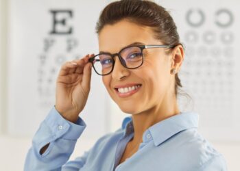 A woman wearing black-rimmed glasses and a blue blouse smiles and touches her glasses in front of a set of eye-test charts.