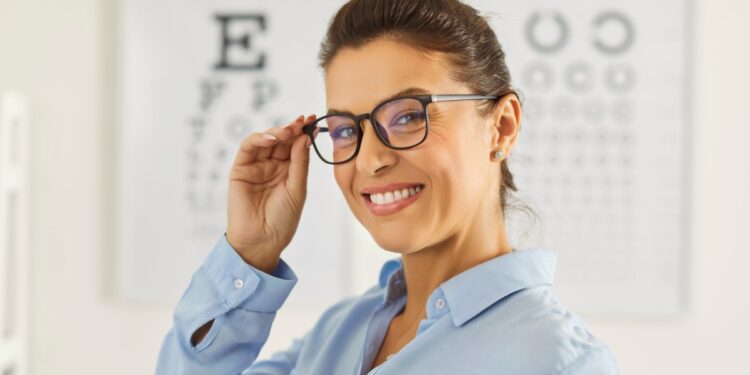 A woman wearing black-rimmed glasses and a blue blouse smiles and touches her glasses in front of a set of eye-test charts.