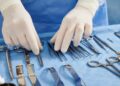 A close-up of a surgeon wearing a blue gown and white gloves, looking at a table of various surgical equipment.
