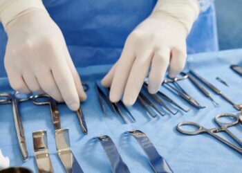 A close-up of a surgeon wearing a blue gown and white gloves, looking at a table of various surgical equipment.