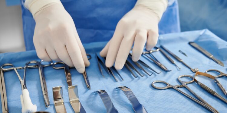A close-up of a surgeon wearing a blue gown and white gloves, looking at a table of various surgical equipment.