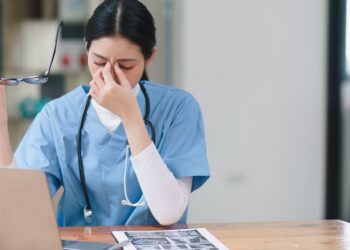 A person wearing scrubs and a stethoscope holds their glasses in one hand and pinches the bridge of their nose with the other.