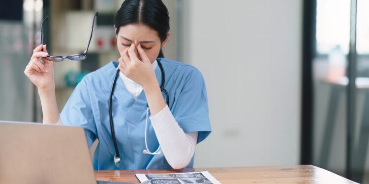 A person wearing scrubs and a stethoscope holds their glasses in one hand and pinches the bridge of their nose with the other.