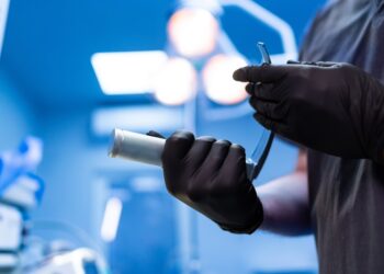A close-up of the hands of a doctor wearing black latex gloves, holding a metal tube inside a hospital room.
