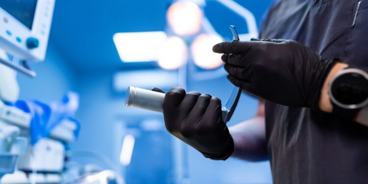 A close-up of the hands of a doctor wearing black latex gloves, holding a metal tube inside a hospital room.