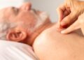A senior man lays down without a top on as a care provider places acupuncture needles into his shoulder.