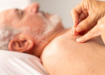 A senior man lays down without a top on as a care provider places acupuncture needles into his shoulder.