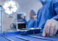 A gloved hand reaches for surgical instruments on a blue drape, with surgeons and lights blurred in the background.