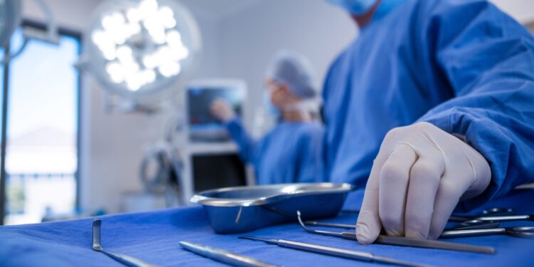 A gloved hand reaches for surgical instruments on a blue drape, with surgeons and lights blurred in the background.
