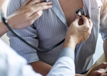 A doctor listens to a male patient’s heart with a stethoscope while he wears an unbuttoned striped shirt.