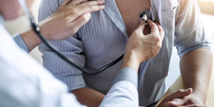 A doctor listens to a male patient’s heart with a stethoscope while he wears an unbuttoned striped shirt.