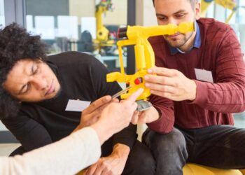 Two men examine a yellow robotic arm apparatus, adjusting components together in a modern lab workspace.