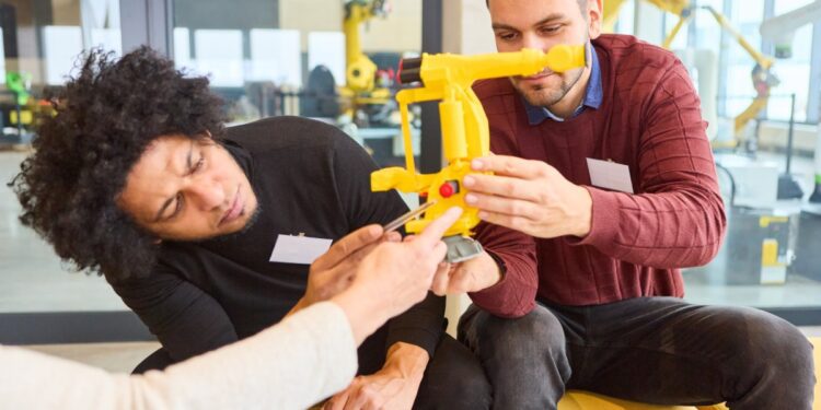 Two men examine a yellow robotic arm apparatus, adjusting components together in a modern lab workspace.