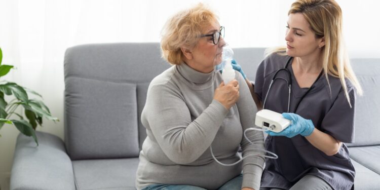 An older woman sits on a couch next to a medical professional. The older woman holds an oxygen mask up to her face.