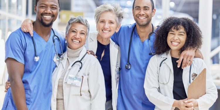 A team of healthcare professionals wearing scrubs and lab coats stands together and smiles in a bright clinic.