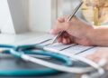 A hand signs a medical form on a desk as a stethoscope rests in the foreground and a globe sits in the background.