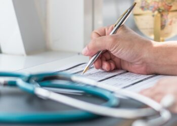A hand signs a medical form on a desk as a stethoscope rests in the foreground and a globe sits in the background.