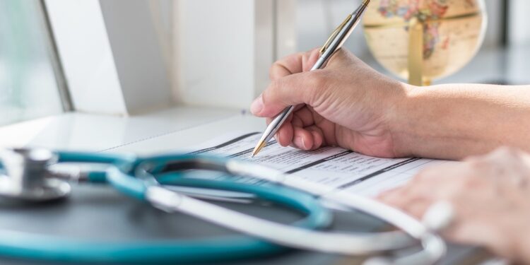 A hand signs a medical form on a desk as a stethoscope rests in the foreground and a globe sits in the background.