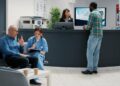 A healthcare waiting room with one doctor talking with a seated patient and another person talking at the reception desk.