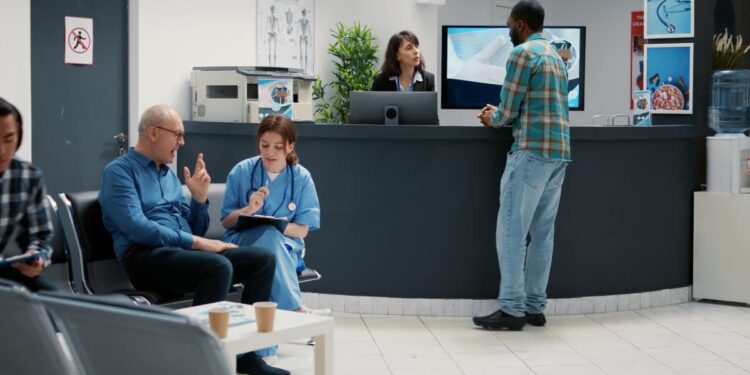 A healthcare waiting room with one doctor talking with a seated patient and another person talking at the reception desk.
