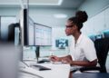 A woman with tied up hair is sitting at a desk and is looking at a computer screen. She is wearing white clothes.