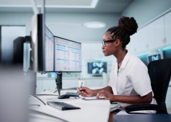 A woman with tied up hair is sitting at a desk and is looking at a computer screen. She is wearing white clothes.