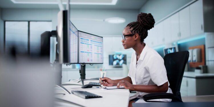 A woman with tied up hair is sitting at a desk and is looking at a computer screen. She is wearing white clothes.
