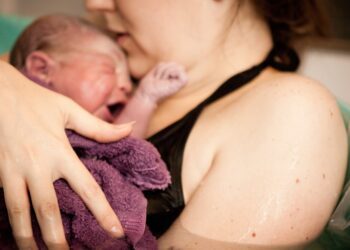 A woman is holding a newborn baby sitting in a birthing pool full of water. A burgundy towel rests on the baby.