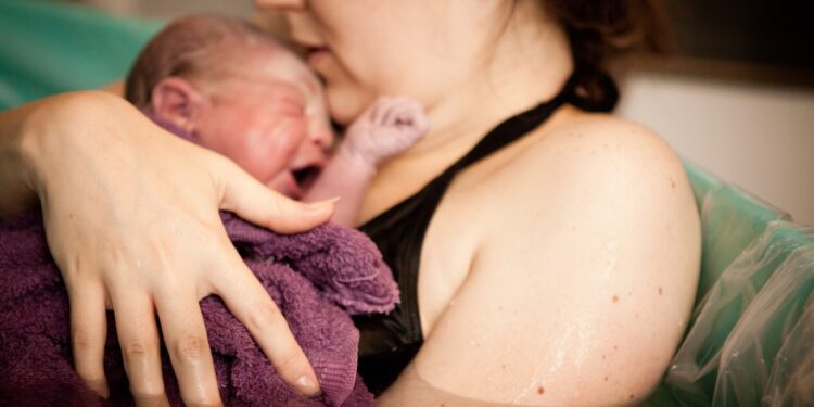 A woman is holding a newborn baby sitting in a birthing pool full of water. A burgundy towel rests on the baby.