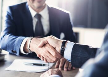 Two men in suits sit across from each other at a table. They are shaking each other's hands above a pile of papers.