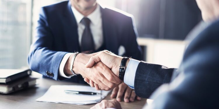 Two men in suits sit across from each other at a table. They are shaking each other's hands above a pile of papers.