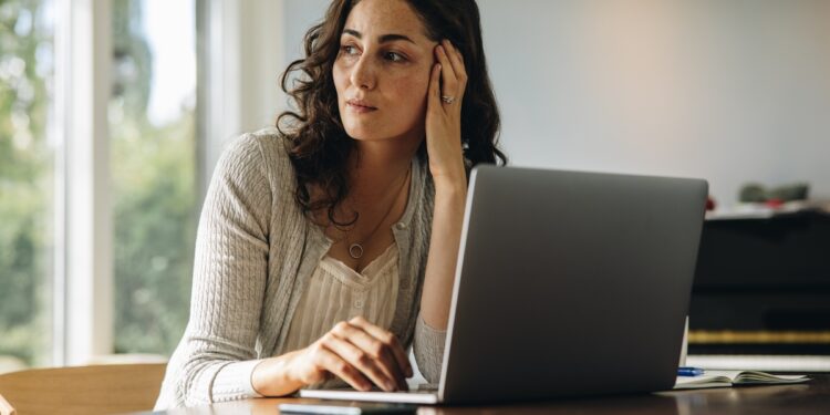 A woman in front of a laptop has one hand on the keyboard and the other pressed against her face. She looks to her side.