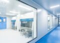 A healthcare facility's long hallway with blue flooring and a large window showing a cleanroom to the left.