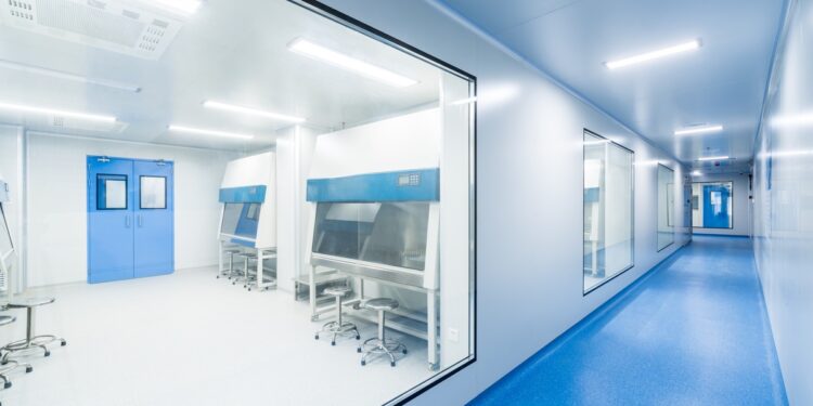 A healthcare facility's long hallway with blue flooring and a large window showing a cleanroom to the left.