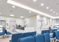 Modern hospital waiting room area with bright, overhead lighting illuminating a room of blue chairs.