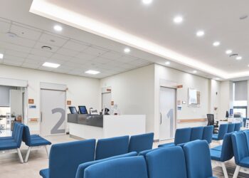 Modern hospital waiting room area with bright, overhead lighting illuminating a room of blue chairs.