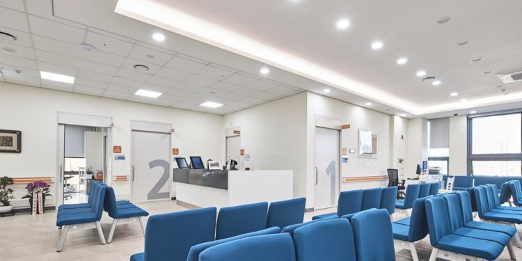 Modern hospital waiting room area with bright, overhead lighting illuminating a room of blue chairs.