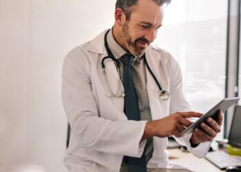 A doctor tapping a tablet near a window in their office during the daytime. The doctor smiles at the tablet.