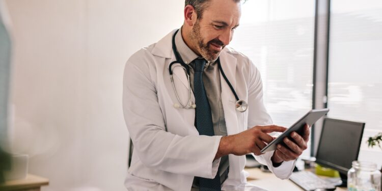 A doctor tapping a tablet near a window in their office during the daytime. The doctor smiles at the tablet.