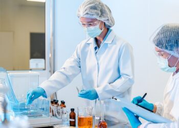 Two scientists wearing white lab coats, gloves, hairnets, face masks, and safety glasses while performing tests in a lab.