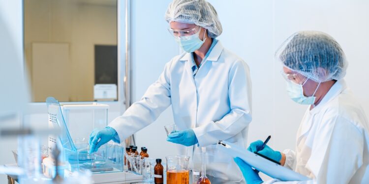 Two scientists wearing white lab coats, gloves, hairnets, face masks, and safety glasses while performing tests in a lab.