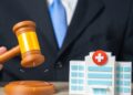 A man wearing a suit raising a judge's gavel beside a small model hospital building on a desk in a bright office.