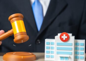A man wearing a suit raising a judge's gavel beside a small model hospital building on a desk in a bright office.