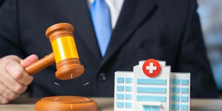 A man wearing a suit raising a judge's gavel beside a small model hospital building on a desk in a bright office.