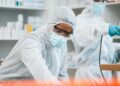 Two workers in protective suits, masks, goggles, and gloves handle equipment in a clean laboratory or pharmacy.