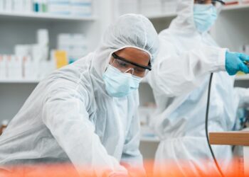 Two workers in protective suits, masks, goggles, and gloves handle equipment in a clean laboratory or pharmacy.