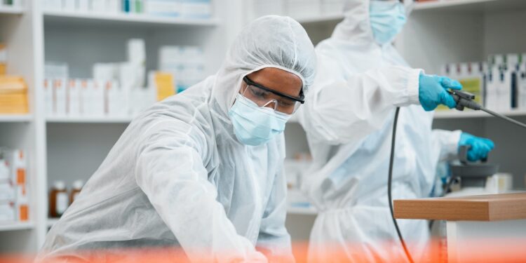 Two workers in protective suits, masks, goggles, and gloves handle equipment in a clean laboratory or pharmacy.