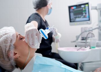 A woman lying in a dental chair while a gloved person holds an anesthesia mask near her face during dental care.